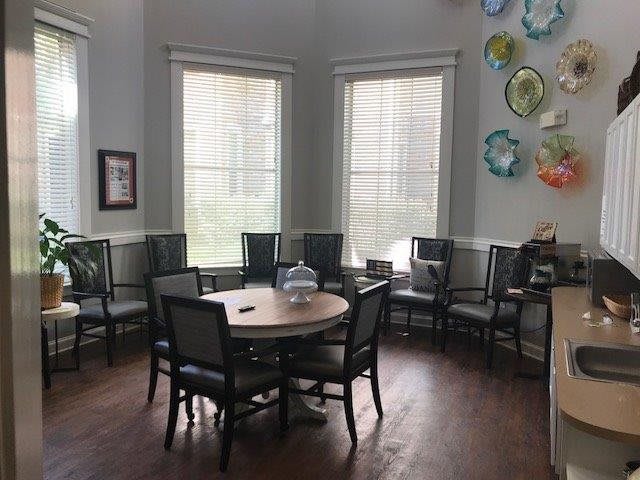A cozy room with a round table surrounded by chairs, illuminated by natural light from large windows and adorned with colorful wall art.