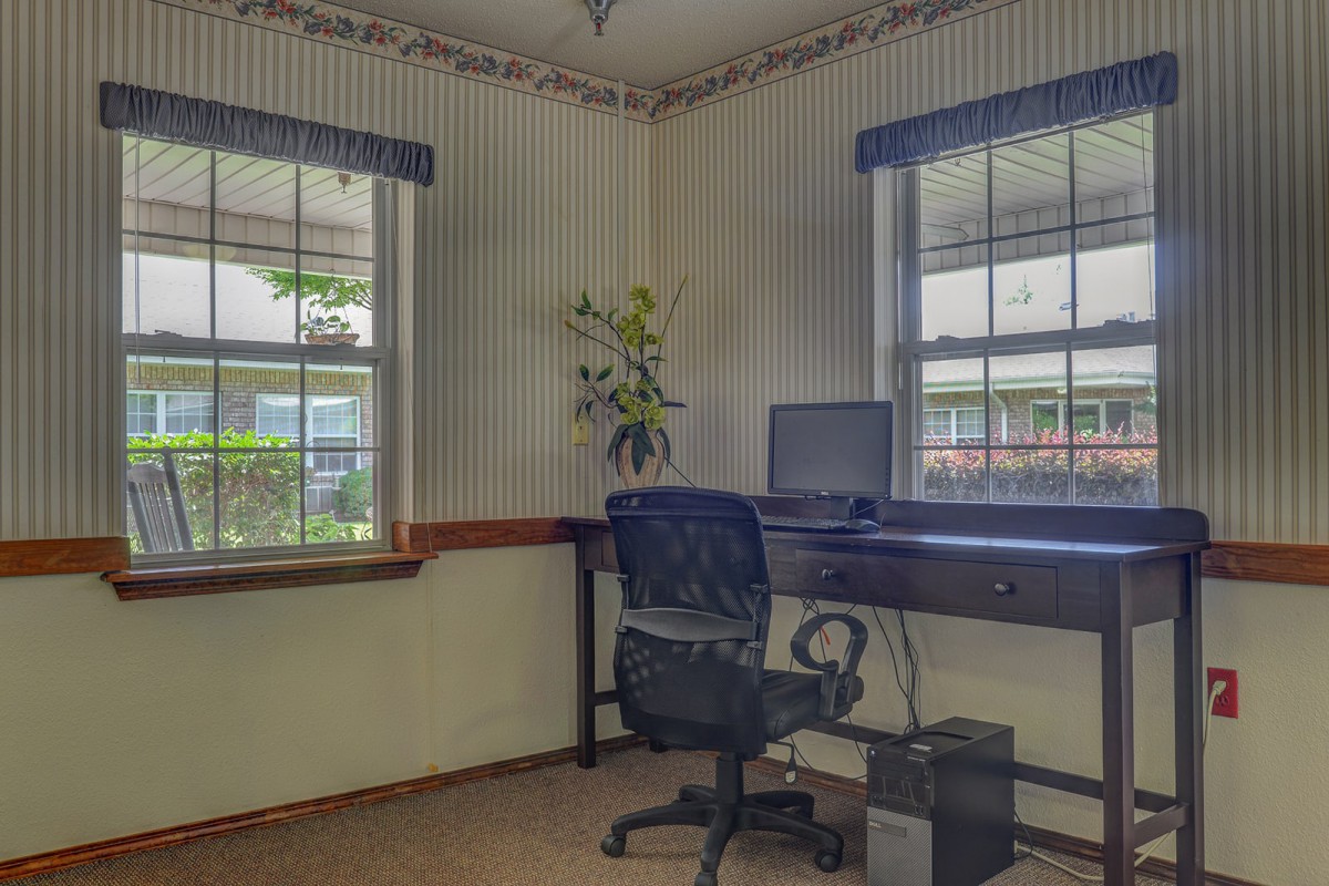 A home office with a desk, computer, and chair near two windows.