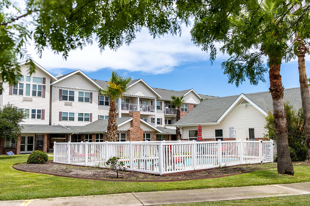 A residential building with a white fence and palm trees under a cloudy sky.