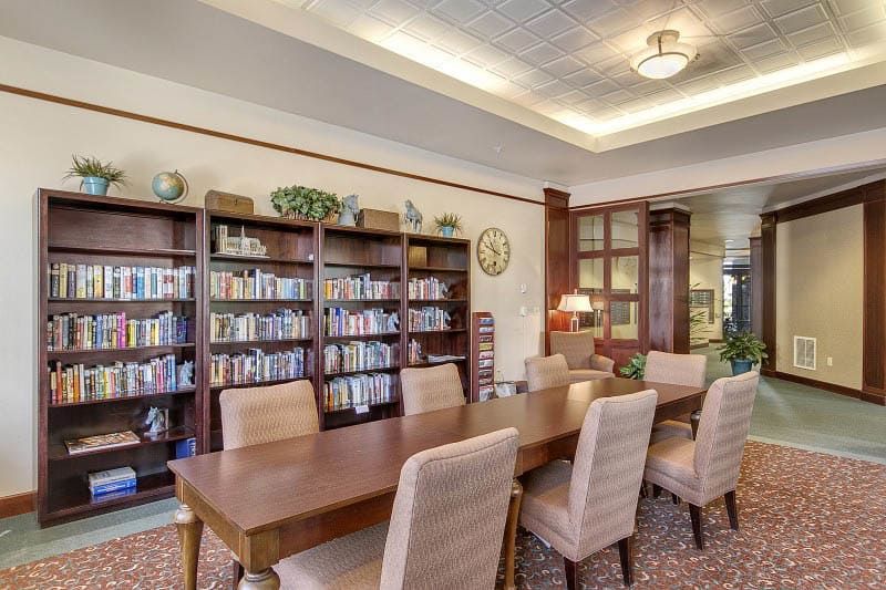 A cozy library room with bookshelves, a large wooden table, and upholstered chairs.