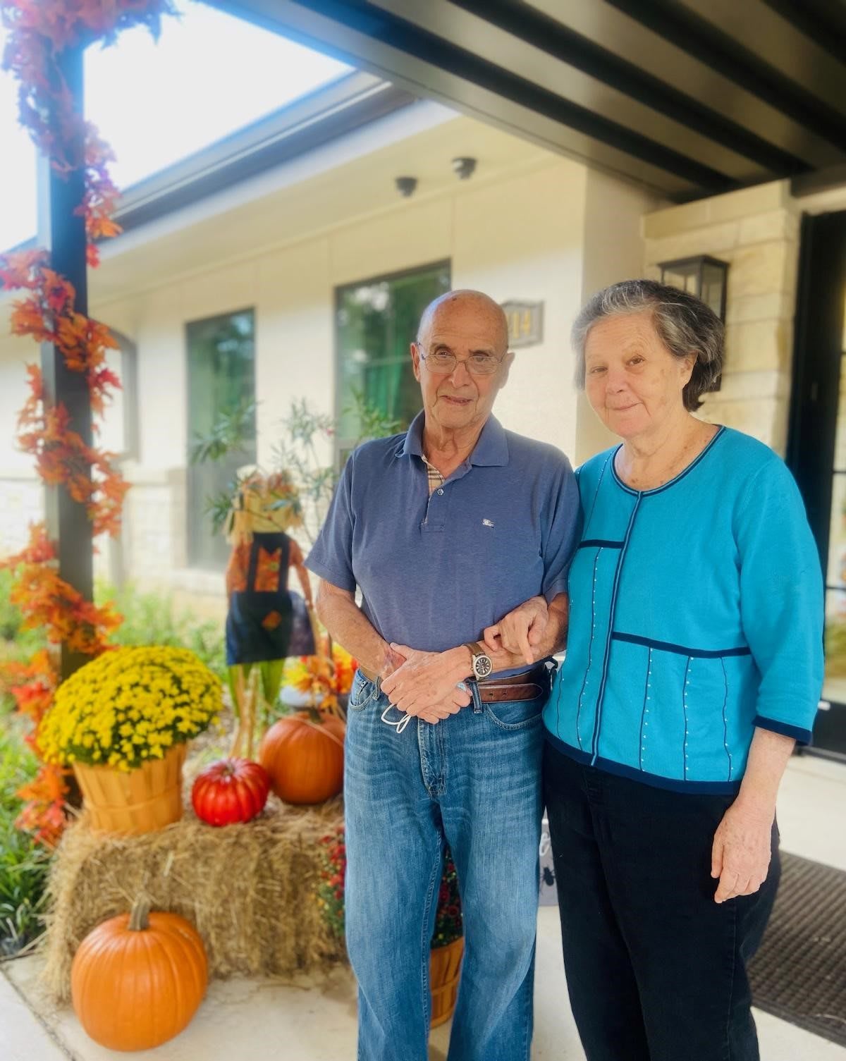An elderly couple stands together in front of a house decorated for autumn with pumpkins and hay bales.