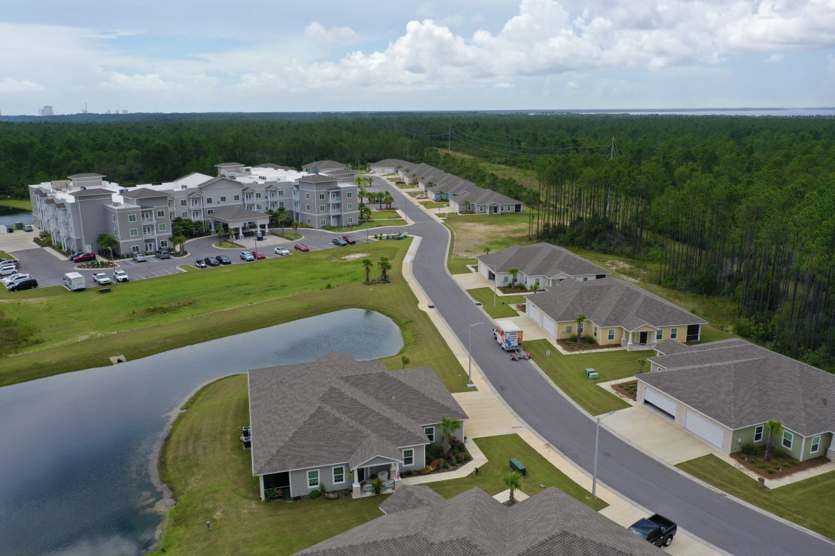 Aerial view of a suburban neighborhood with houses, a road, and a pond surrounded by trees.