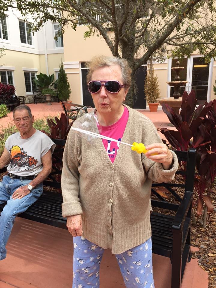 An elderly woman wearing sunglasses blows bubbles while standing near a bench outdoors.
