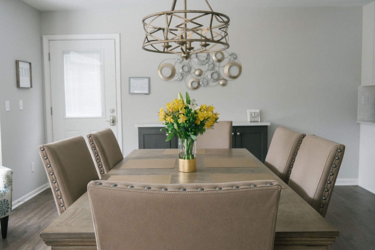 A modern dining room featuring a wooden table with beige upholstered chairs, a stylish chandelier overhead, and a vibrant bouquet of yellow flowers as the centerpiece.