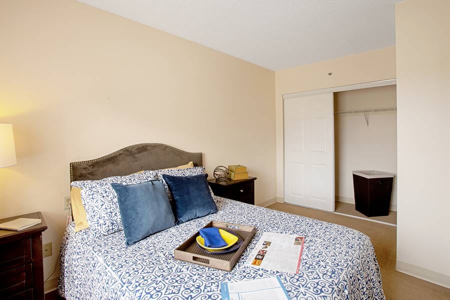 A cozy bedroom with a neatly made bed, featuring decorative pillows and a tray on top. There's a dresser beside the bed and an open closet in the background.