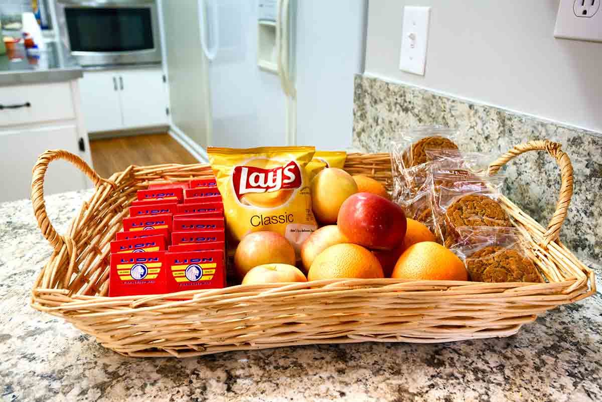 A basket on a kitchen counter filled with assorted snacks, fruits, and granola bars.