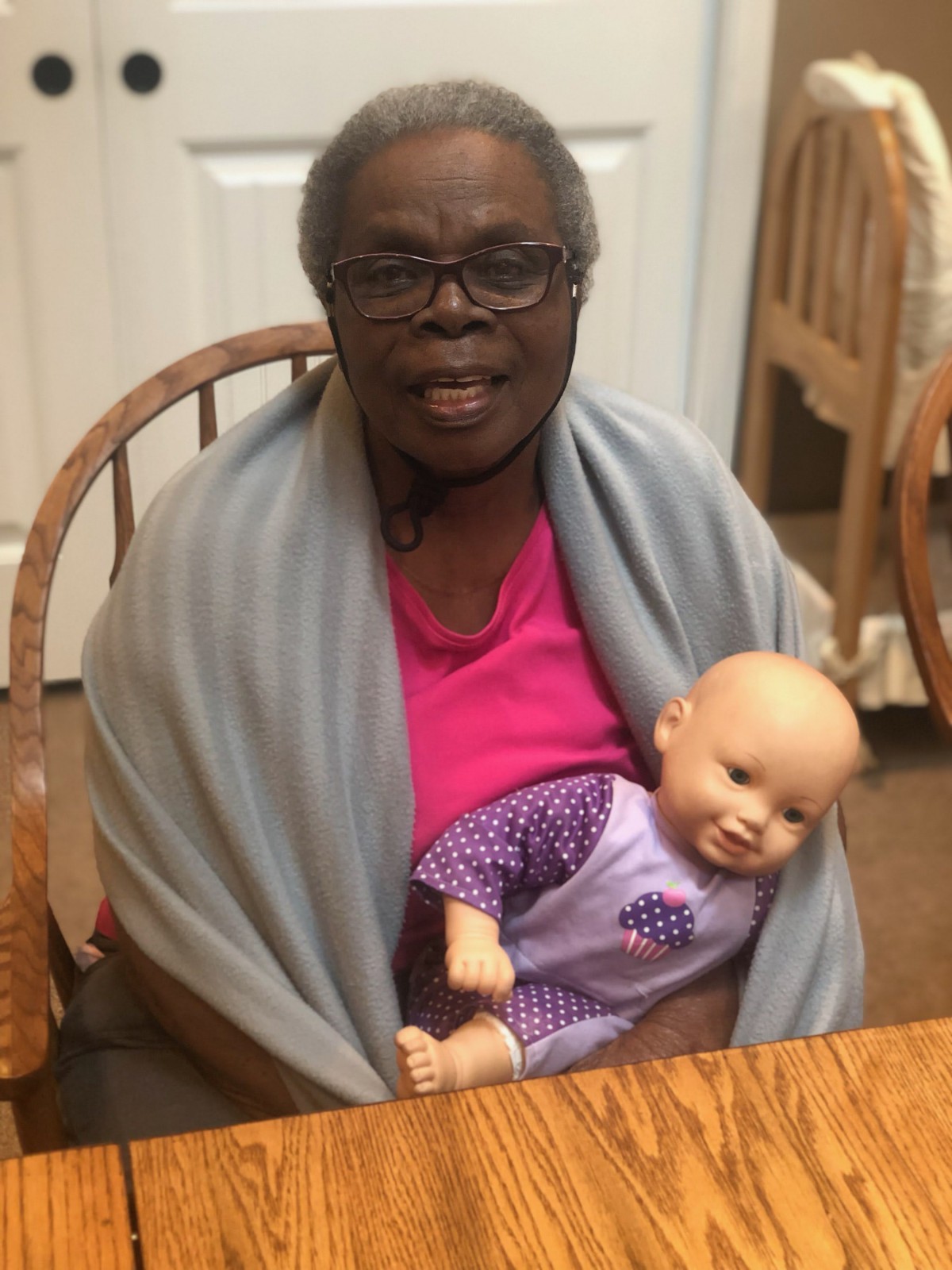 A woman with glasses sitting at a table, smiling and holding a doll wrapped in a shawl.