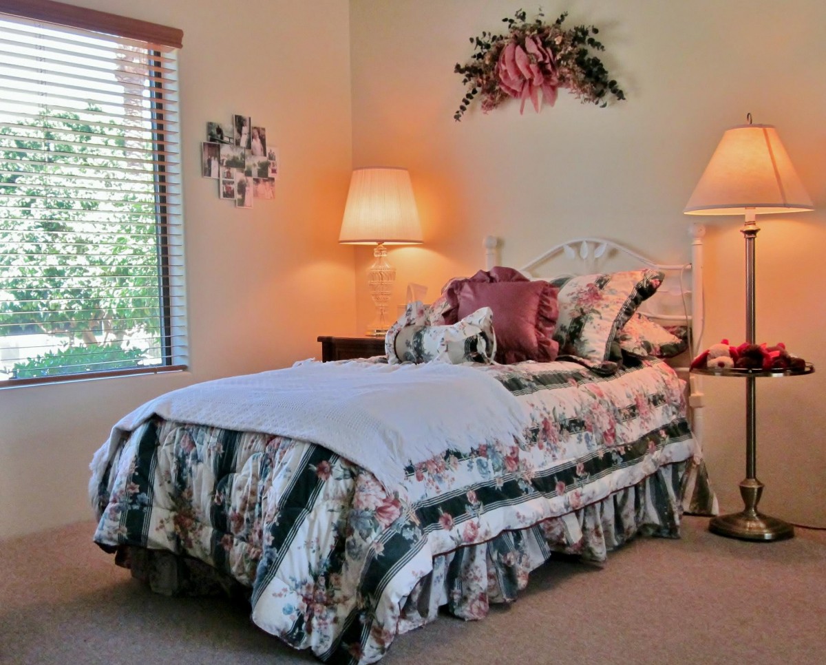 A cozy bedroom featuring floral bedding, warm lighting from two lamps, and a window with blinds.