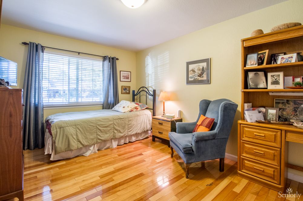 A cozy bedroom with a single bed, armchair, and bookshelf, featuring wooden floors and a large window.