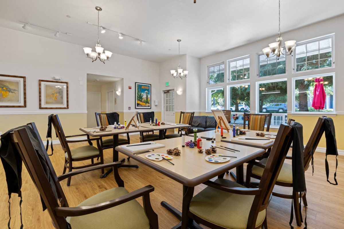 A well-lit dining room with neatly arranged tables and chairs, featuring large windows and elegant chandeliers.