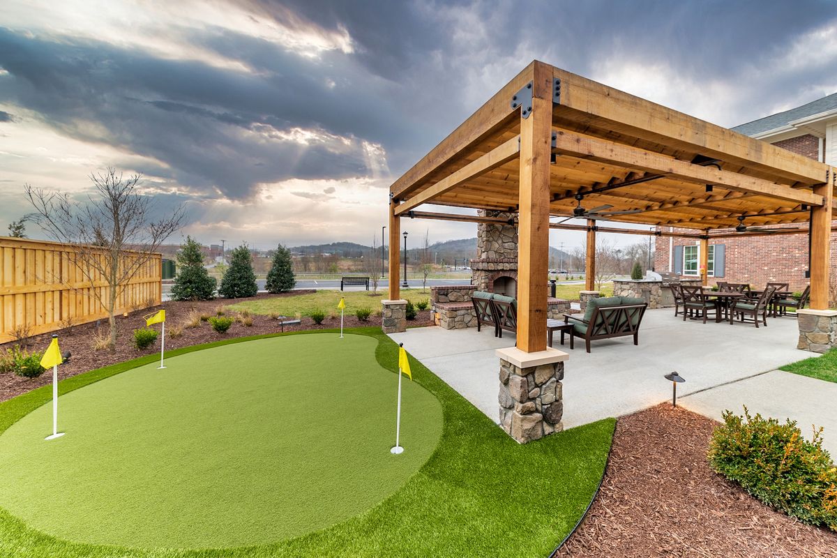 A beautiful outdoor patio with a pergola, featuring a cozy seating area, fireplace, and an adjacent putting green under a cloudy sky.