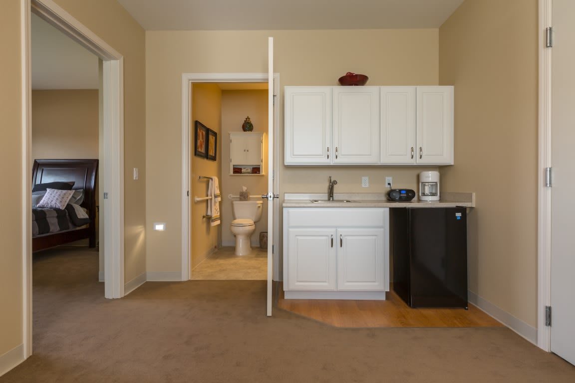 A small kitchenette with white cabinets, a mini fridge, and a microwave next to a bathroom entrance.