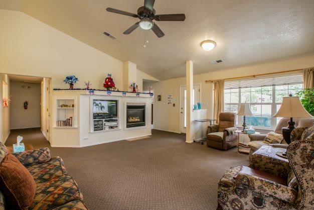 A cozy living room featuring a fireplace, ceiling fan, armchairs, and large windows.