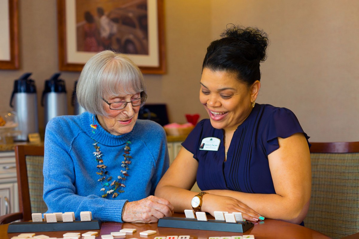 A caregiver assisting an elderly woman with a board game, both smiling.