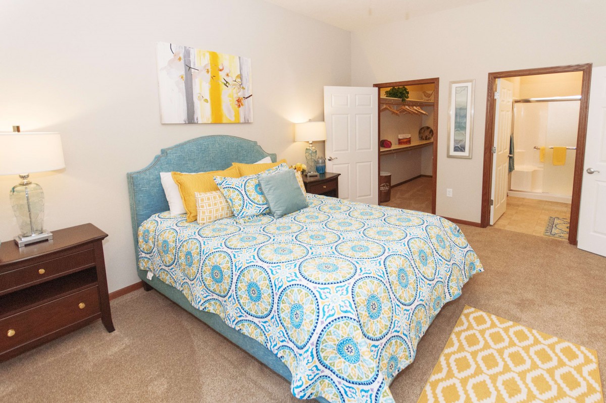 A cozy bedroom with a bed featuring blue and yellow bedding, complemented by a small open closet and decorative wall art.