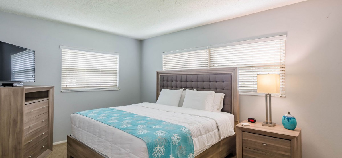 A cozy bedroom featuring a large bed with white and turquoise bedding, illuminated by natural light from two windows with blinds.