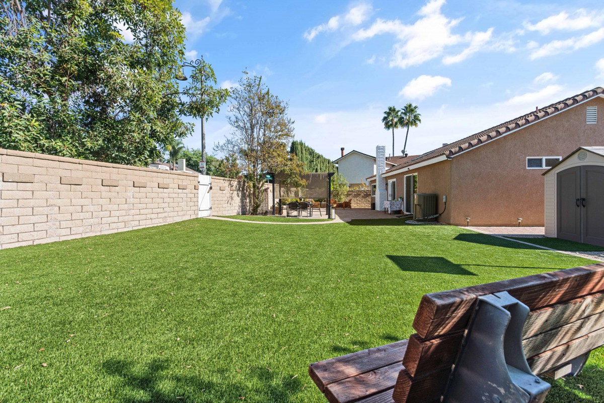A sunny backyard featuring a well-maintained lawn, surrounded by trees and a fence, with a bench and a house in the background.