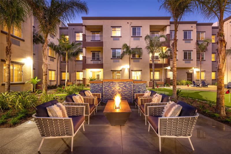 A cozy outdoor seating area with a fire pit in the courtyard of an apartment complex, highlighted by palm trees and evening lighting.