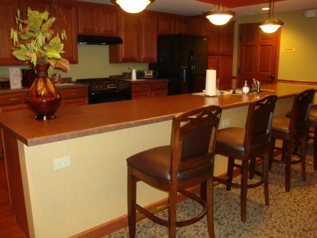 A cozy kitchen with wooden cabinets and a long countertop, featuring bar stools and decorative lighting.