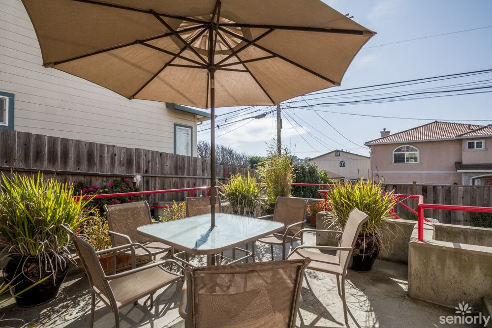 A sunny outdoor patio with a table, chairs, and an umbrella surrounded by potted plants.