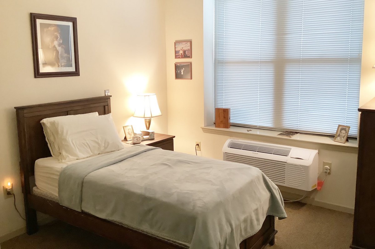 A cozy bedroom with a single bed, nightstand, and lamp next to a large window.