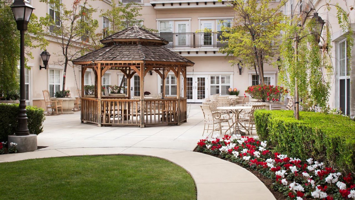 A scenic outdoor garden featuring a wooden gazebo surrounded by colorful flowers and a well-maintained lawn.