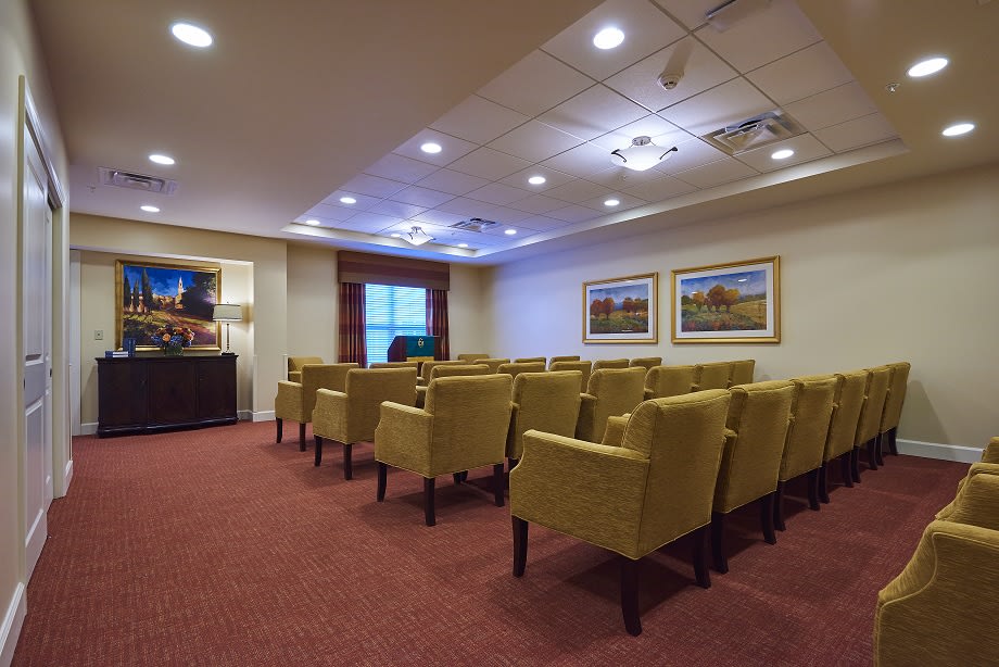 A well-lit conference room with rows of yellow upholstered chairs facing a screen, featuring carpet flooring and decorative paintings on the walls.