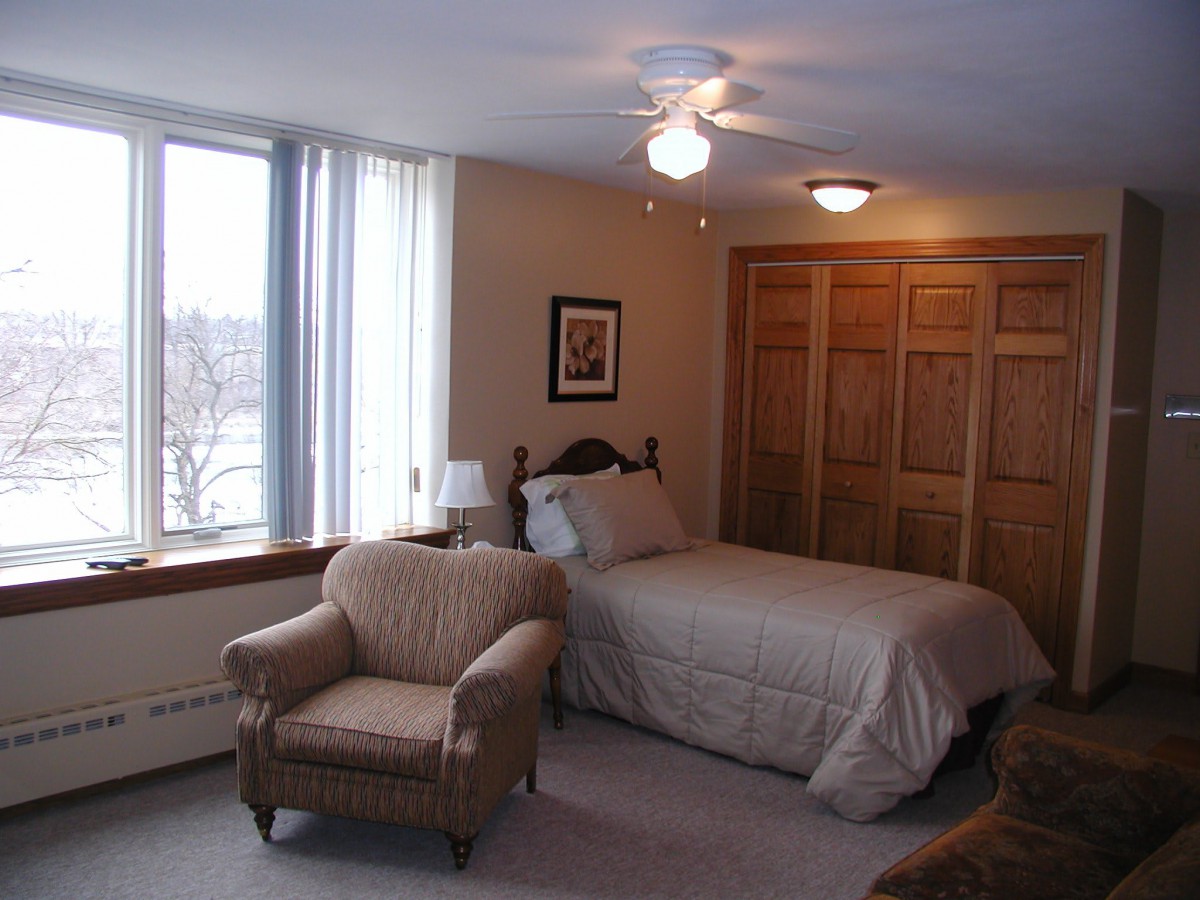 A cozy bedroom with a bed, armchair, and large window letting in natural light.