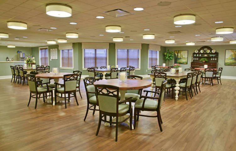 A spacious dining room with multiple tables and chairs, featuring soft lighting and large windows.
