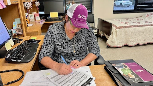 A woman in an office setting is focused on completing paperwork while wearing a purple cap.