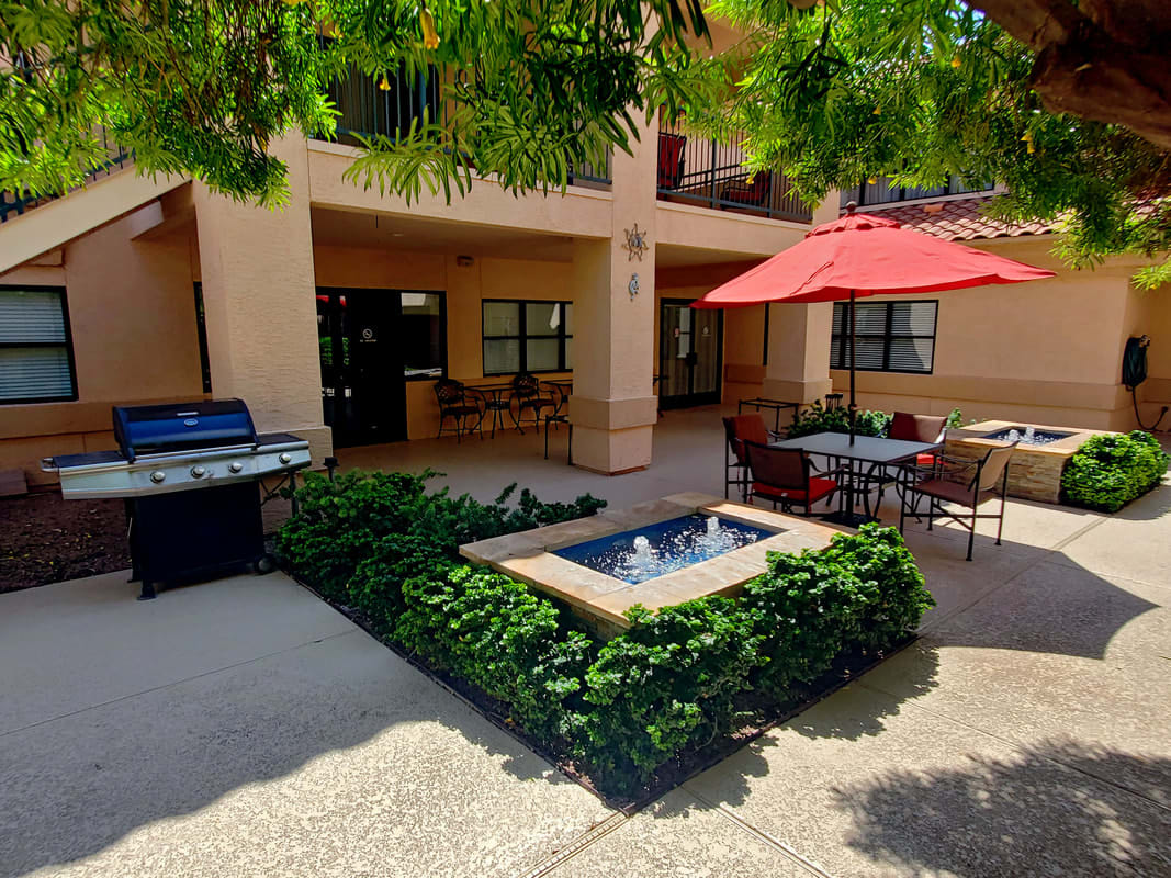 A peaceful courtyard featuring a small fountain, outdoor seating with a red umbrella, and a grill area surrounded by lush greenery.