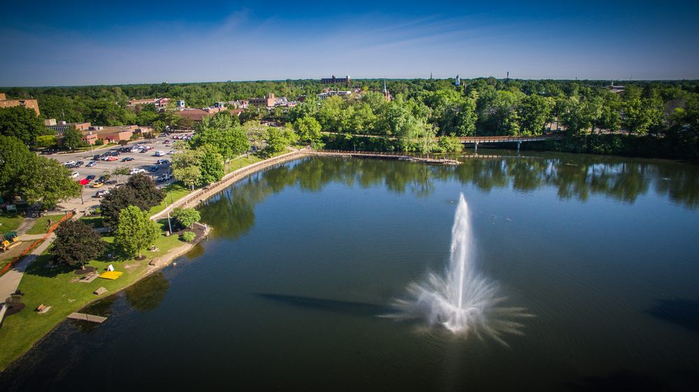 Aerial view of a park featuring a large lake with a prominent fountain at its center, surrounded by lush greenery and city buildings in the background.