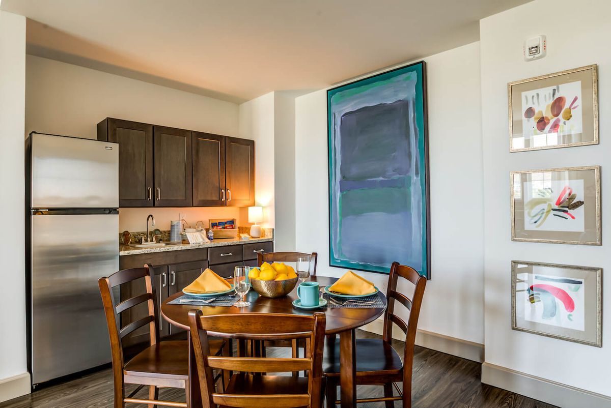 A cozy kitchen and dining area featuring a set wooden table with matching chairs, adorned with stylish tableware and centered by a colorful modern painting.