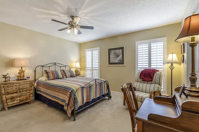 A cozy bedroom featuring a king-sized bed with striped bedding, a ceiling fan, an armchair, and a desk. The room is well-lit with natural light from windows adorned with shutters.