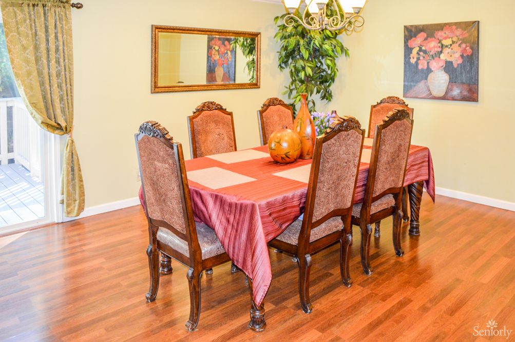 A warmly decorated dining room featuring a wooden dining table with a red tablecloth, surrounded by six ornate chairs. A chandelier hangs overhead and a mirror is mounted on the wall.