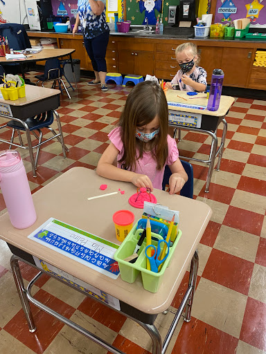 A young girl engages in a creative arts and crafts activity at her school desk, wearing a mask and surrounded by various art supplies.