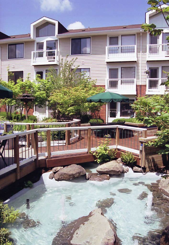 A serene residential courtyard featuring a pond, lush greenery, and a wooden deck with outdoor seating.