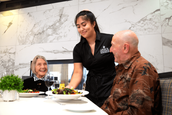 A waitress serves a meal to two seniors in a restaurant setting.