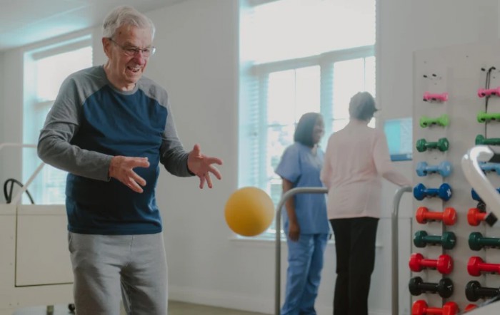 An elderly man engages in physical therapy with a ball under the guidance of healthcare professionals.