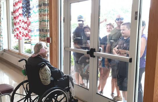 A group of people joyfully interacting with an elderly man in a wheelchair through a glass door.