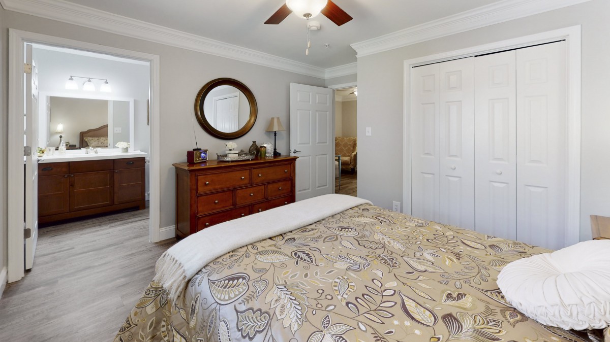 A cozy bedroom featuring a wooden dresser with a round mirror, a bed with patterned bedding, and a ceiling fan.