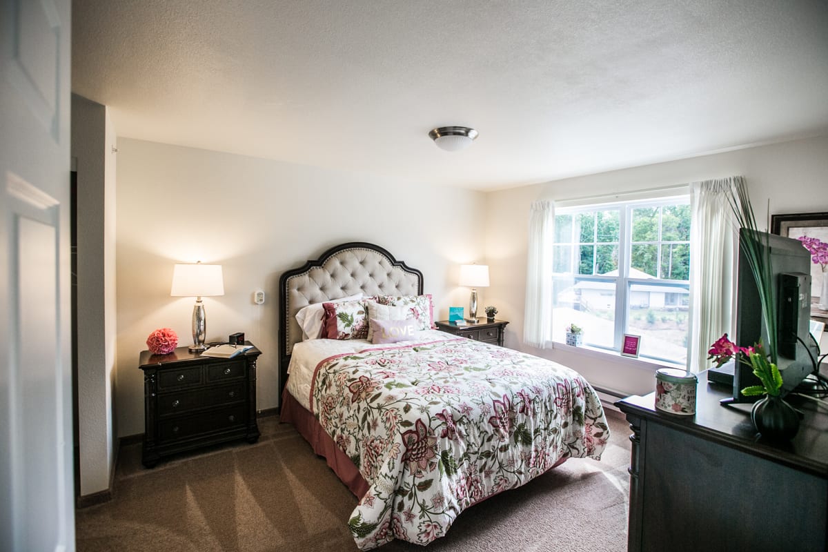 A cozy bedroom with a floral-patterned bedspread, bedside lamps, and a large window letting in natural light.