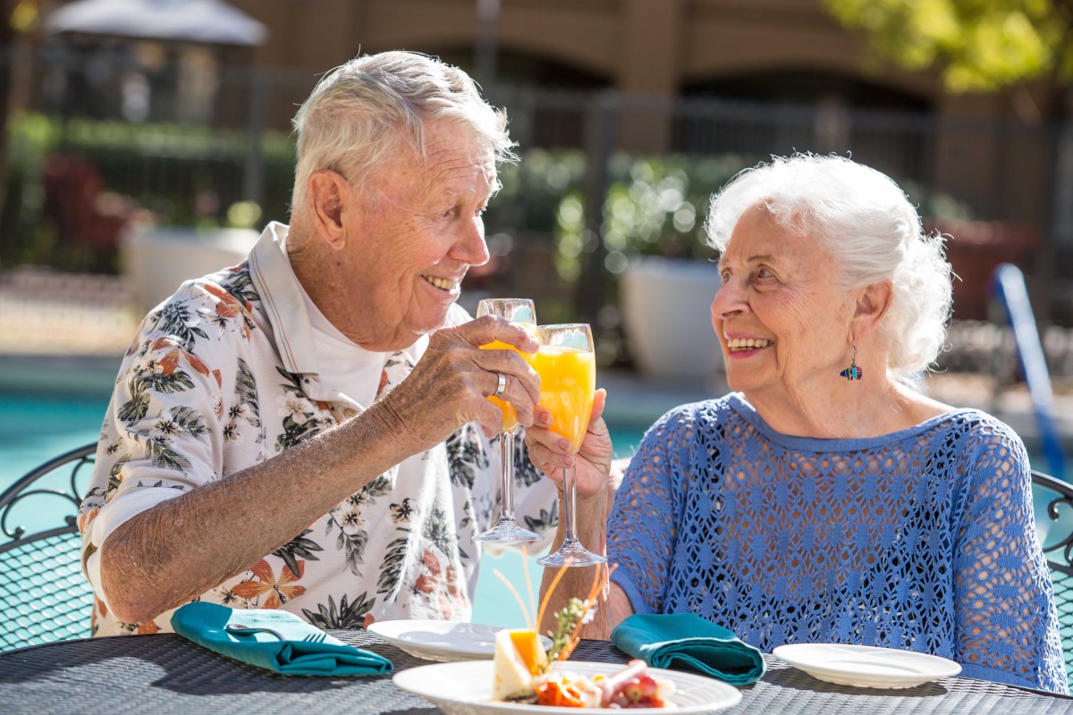 An elderly couple shares a joyful toast during an outdoor brunch.