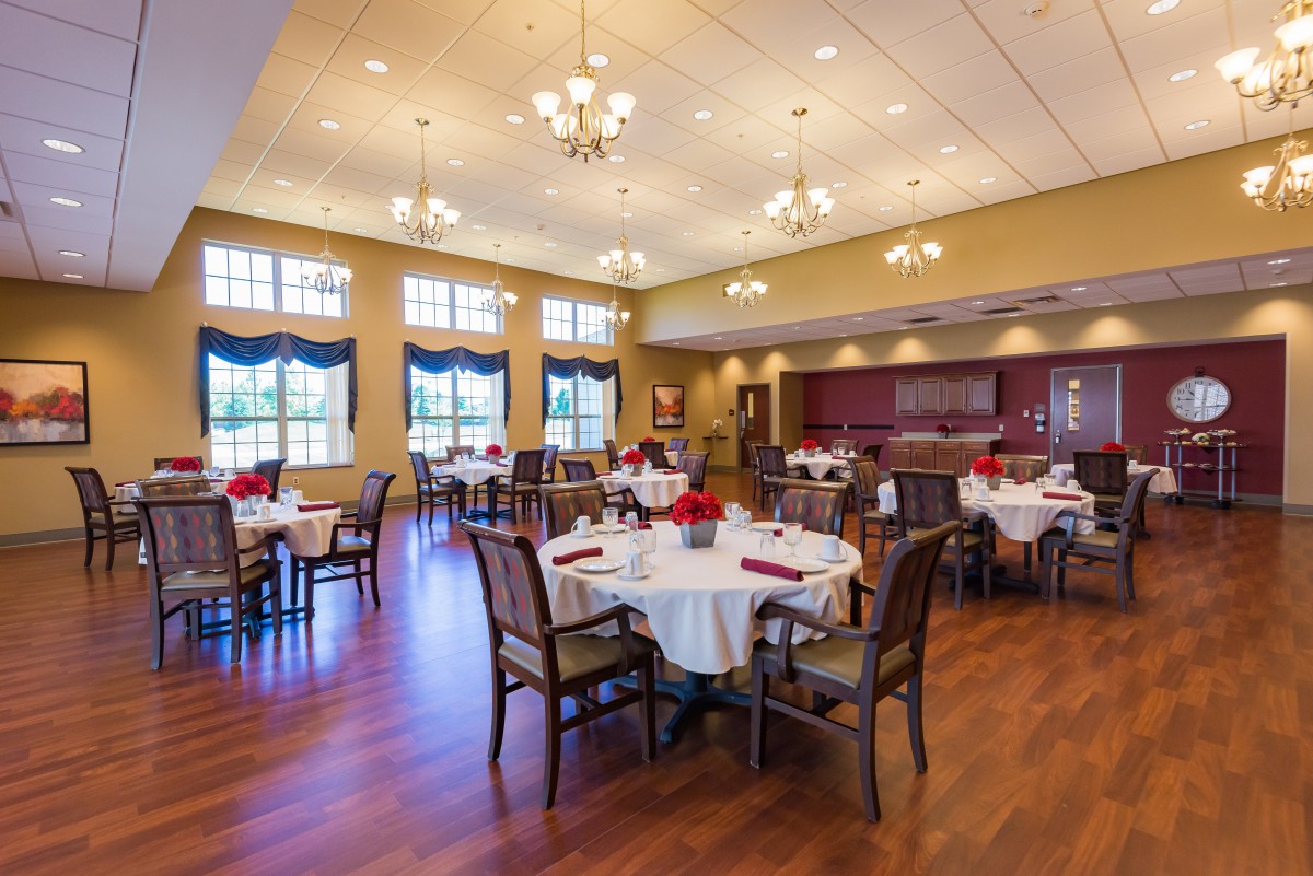 A well-lit dining room with elegantly set tables and chairs, featuring large windows and chandeliers.
