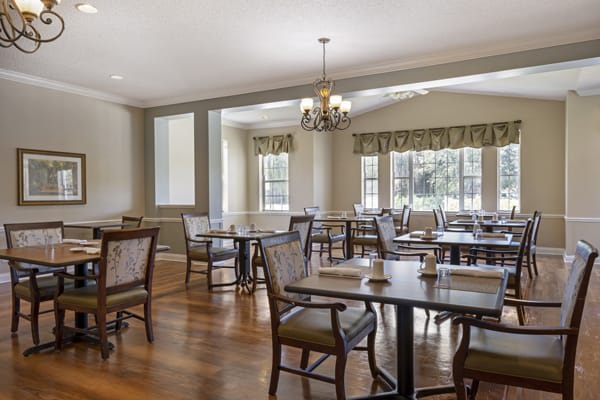 A well-lit dining area with tables and chairs, featuring a chandelier and large windows.