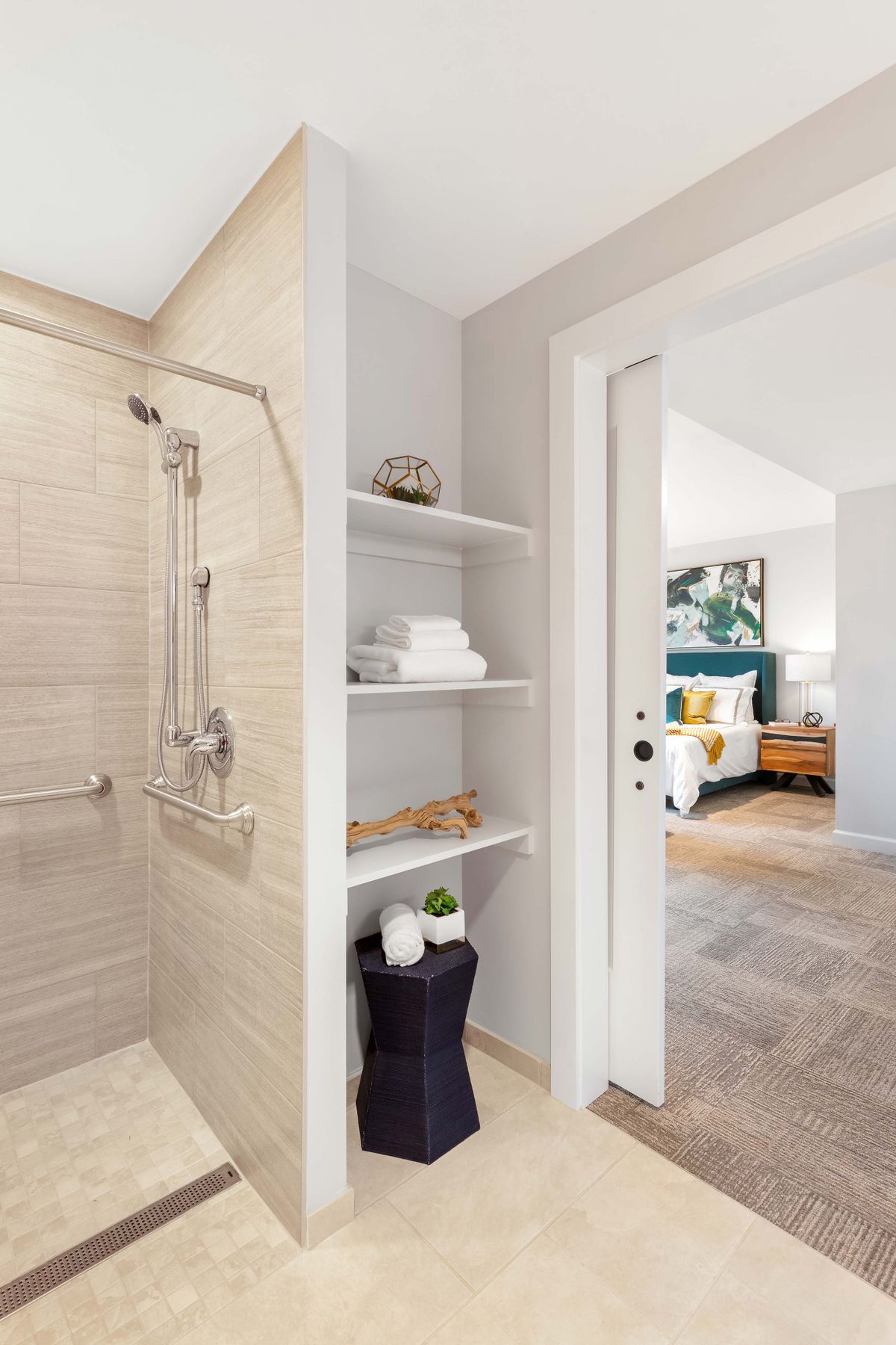 A modern bathroom featuring a walk-in shower and neatly arranged shelves with towels, leading into a cozy bedroom.