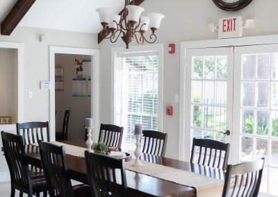 A bright dining room with a long table, chairs, and a chandelier, illuminated by natural light from the windows.