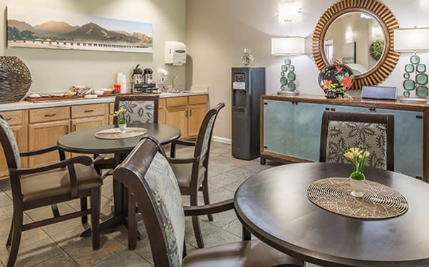 A cozy dining area with neatly arranged tables and chairs, warm lighting, decorative artwork, and a beverage station.