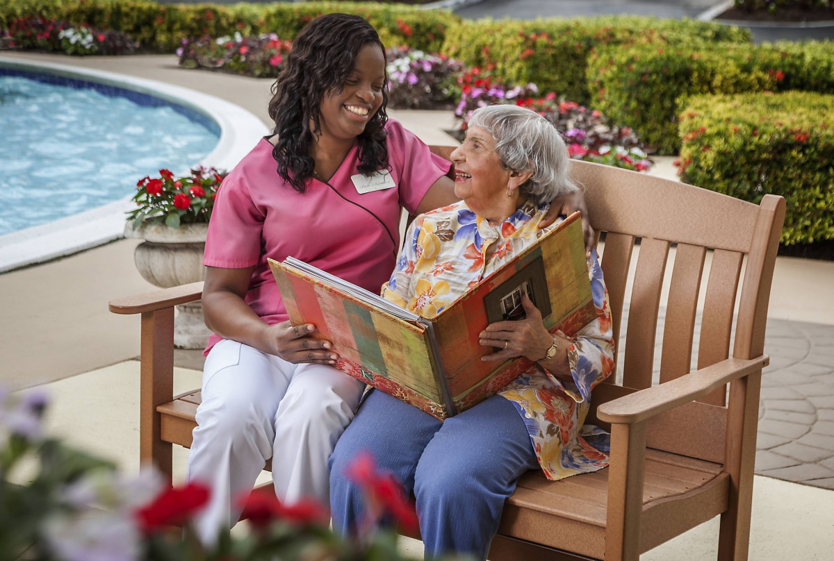 A caregiver and an elderly woman share a book together while sitting on a bench in the garden.