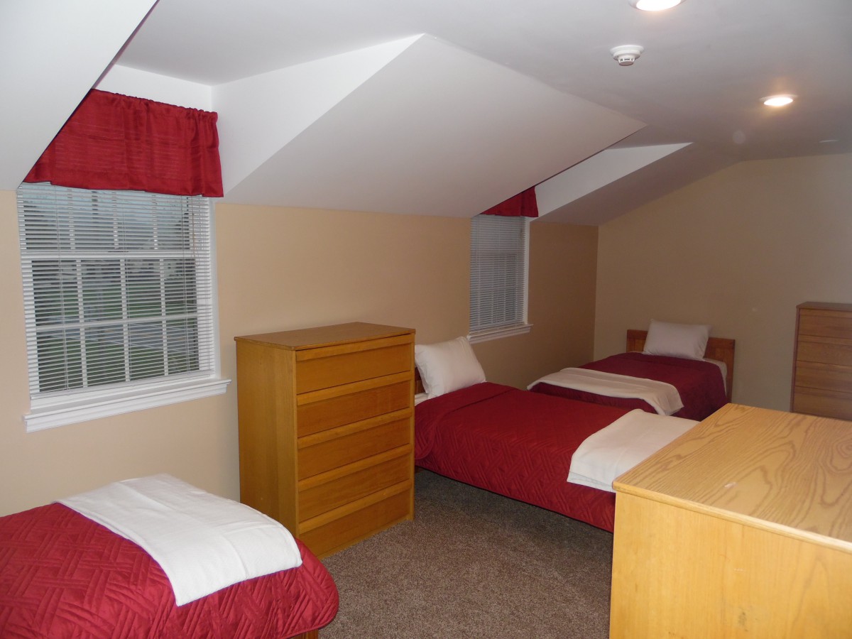 A cozy attic bedroom with twin beds and red bedspreads, featuring a wooden dresser and windows providing natural light.